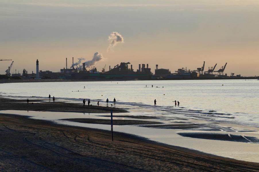 Blick auf die Küste, am Horizont sind Industrieanlagen und aufsteigender Rauch zu sehen.