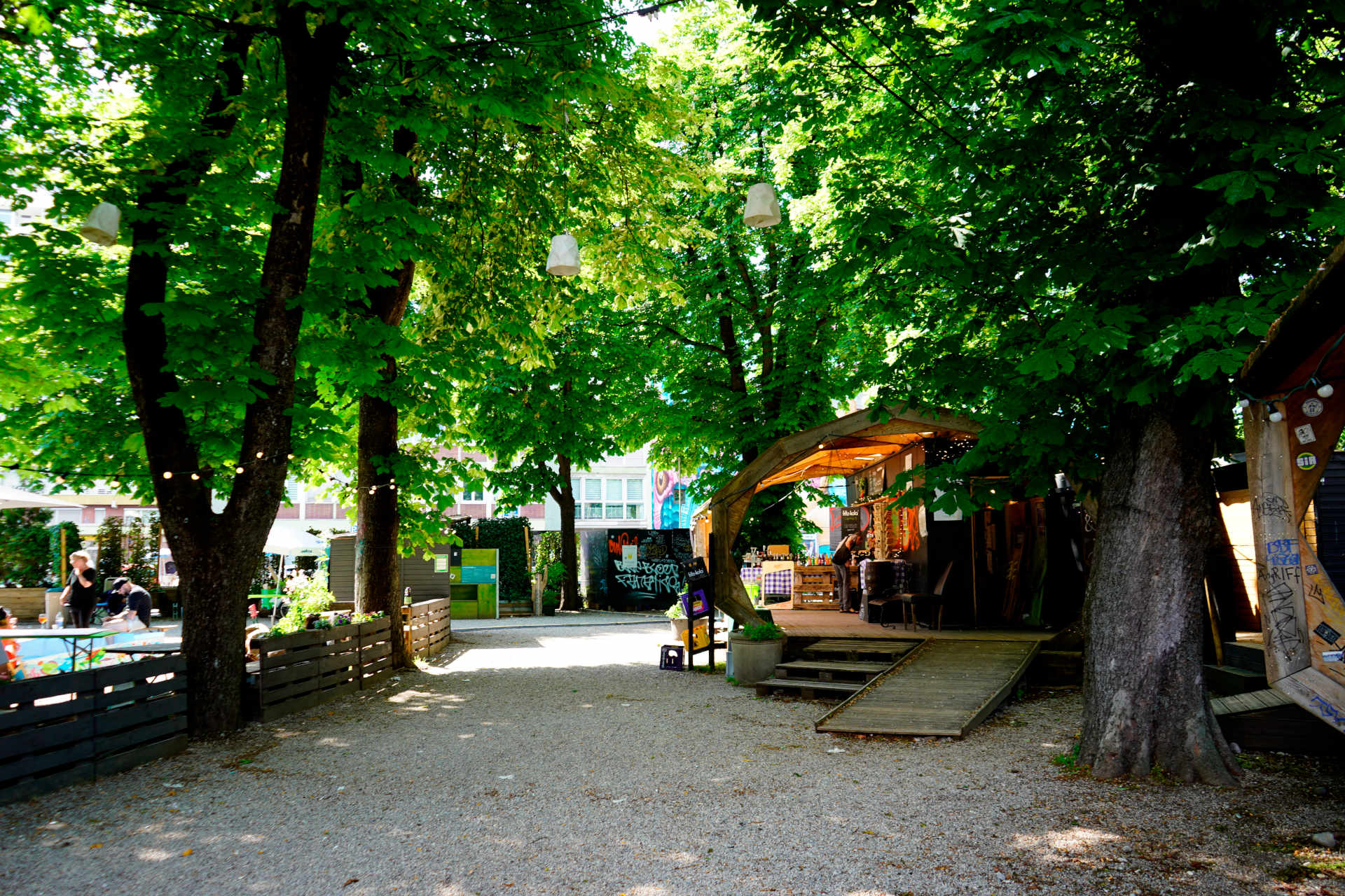 Ein Platz mit großen Kastanienbäumen, darunter Kies, hinten rechts im Bild ein Holzpavillon mit einem Kiosk.