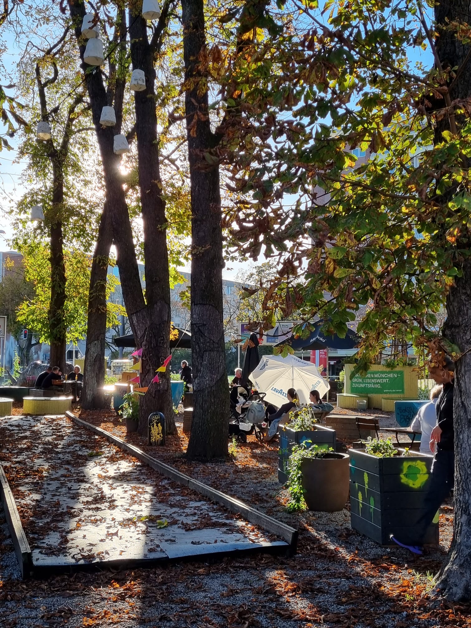 Ein Platz mit großen Kastanienbäumen, darunter links ein mit Blättern bedeckter Bouleplatz, rechts Hochbeete und Menschen, die an Gartentischen sitzen.