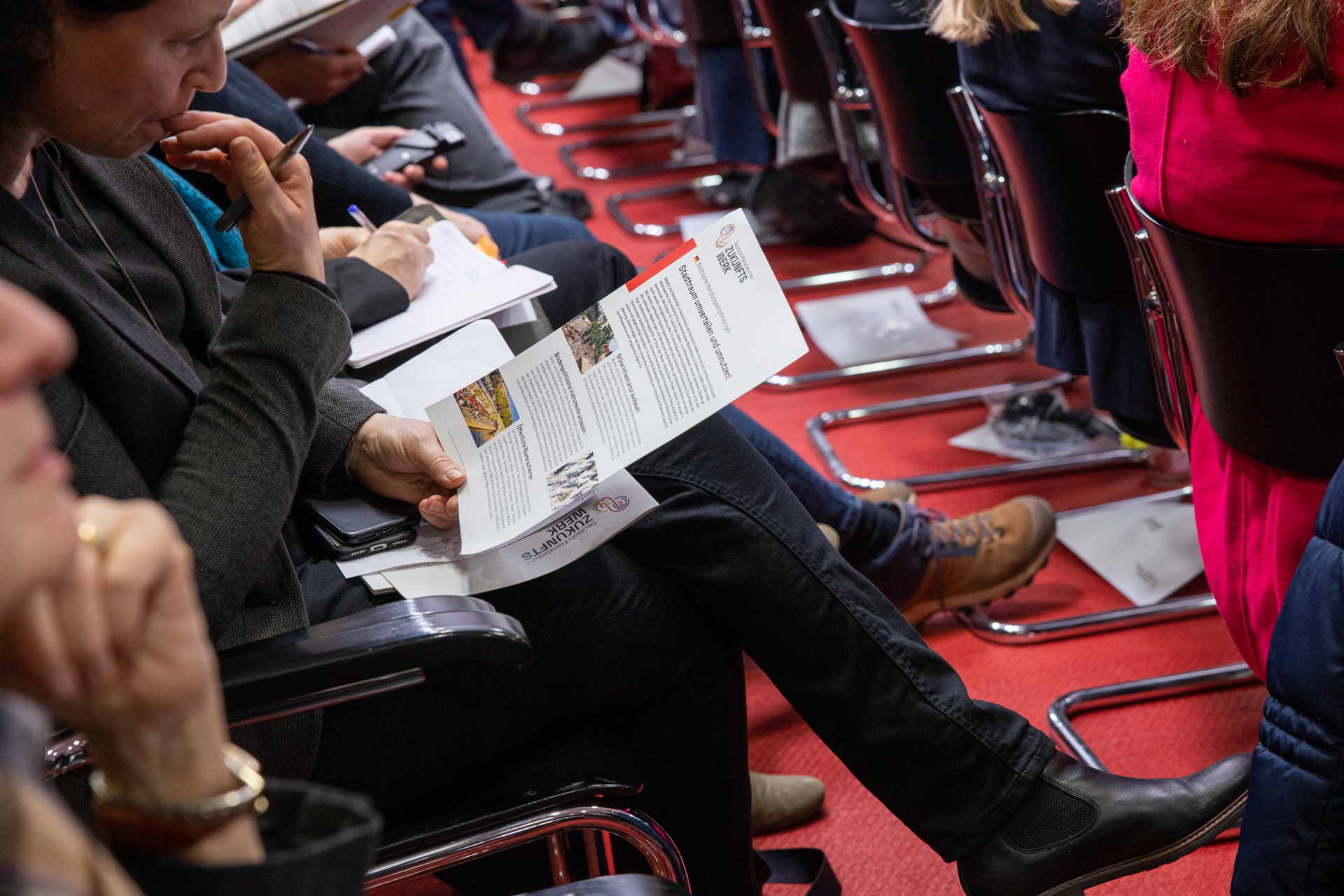 Eine Person sitzt in einem Konferenzsaal und hält ein Dokument in der Hand.