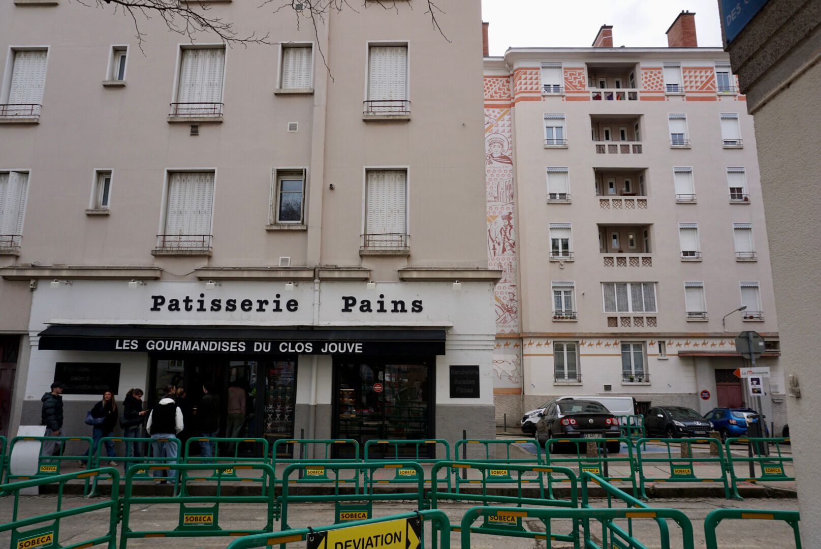 Mehrstöckige Wohngebäude mit Erdgeschoss-Läden, darunter eine Bäckerei mit der Aufschrift "Patisserie Pains", hinter einer grünen Absperrung