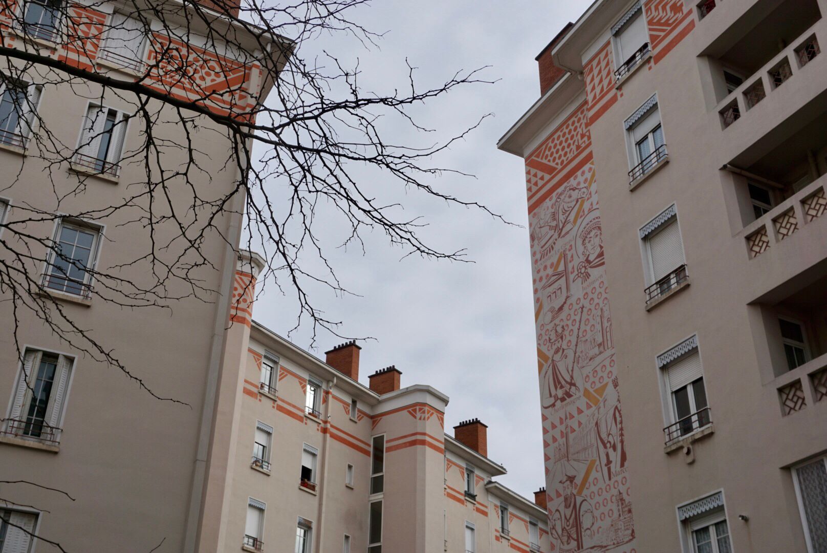 Mehrstöckige, hell verputzte Wohnhäuser mit orangeroten Verzierungen und einem großen, detailreichen Wandgemälde an der Fassade, sind umrahmt von einem kahlen Baum