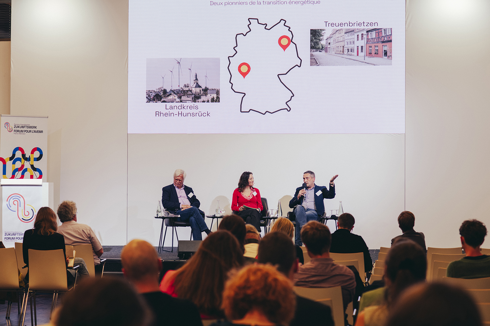 3 Personen diskutieren auf der Bühne während einer Podiumsdiskussion vor einem Publikum