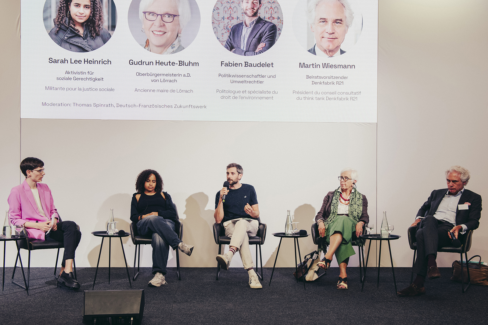 Fünf Personen stehen bei einer Podiumsdiskussion auf einer Bühne, einer von ihnen spricht ins Mikrofon
