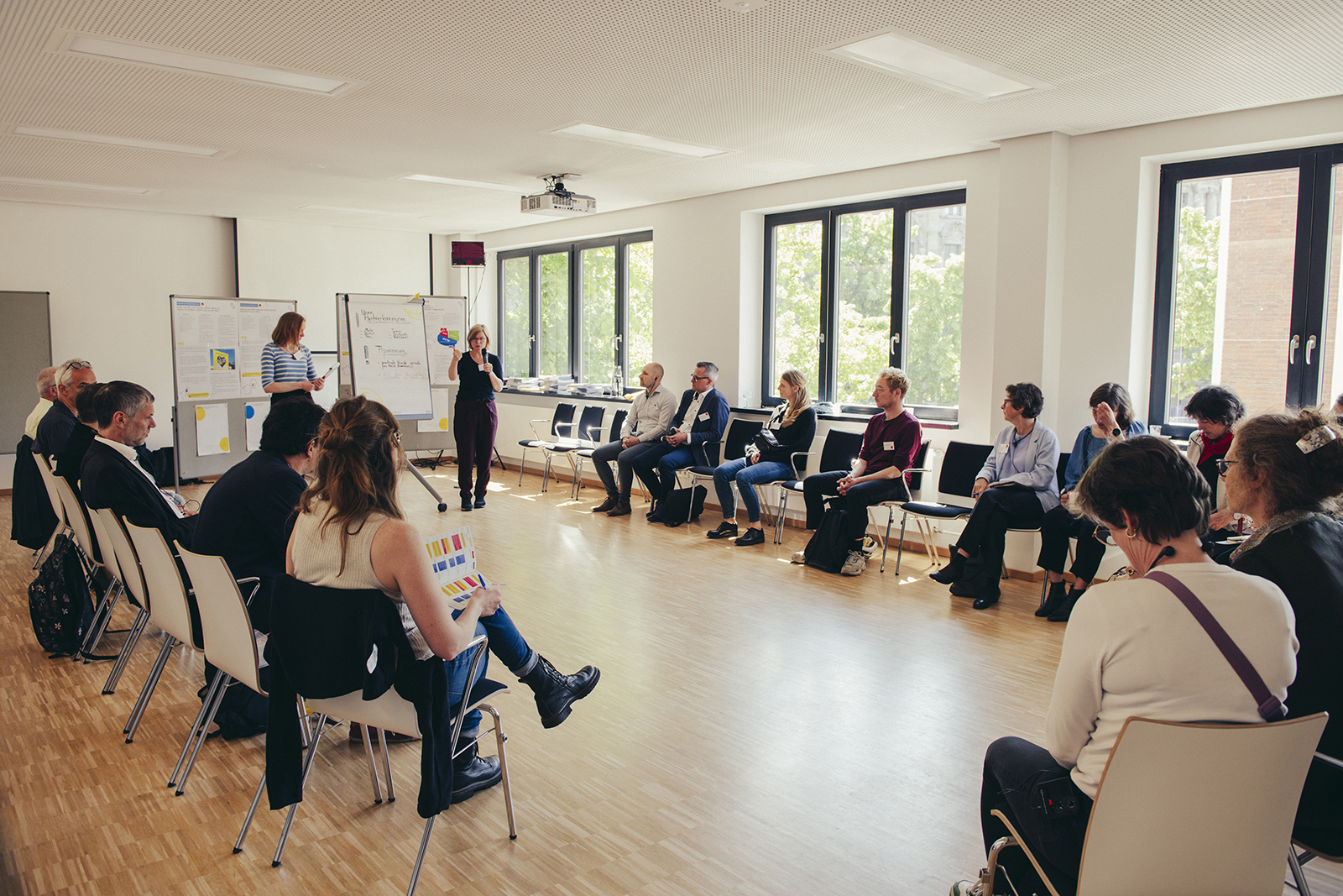 Teilnehmende eines Workshops sitzen in einem Stuhlkreis, vorne wird ein Vortrag gehalten