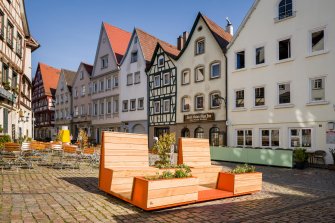 Sesselartige Sitzmöglichkeiten aus Holz inmitten eines von Fachwerkhäusern gesäumten Platzes