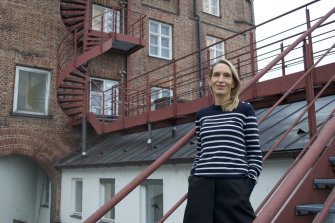 Eine weiblich gelesene Person mit blonden schulterlangen Haaren und schwarz-weiß gestreiftem Pulli steht vor einem Gebäude auf einer Treppe.