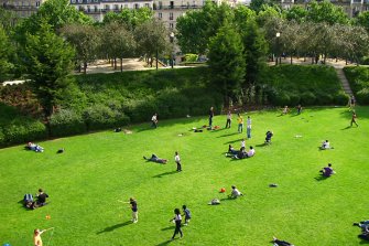 Des personnes sont allongées et marchent sur une pelouse entourée d'arbres. Derrière, on voit des bâtiments.