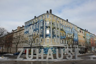 Eine Straße mit kunstvoll bemalten Gebäudefassaden im Hintergrund. Auf der Straße steht der Schriftzug „Zuhause“ in riesigen Lettern aus lackiertem Stahlblech.