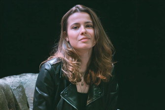 Une personne aux longs cheveux bruns et portant une veste en cuir noire est assise dans un fauteuil gris.
