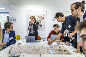 Menschen sitzen um einen Tisch und notieren auf einem A1-Plakat. Eine Person spricht in ein Mikro.