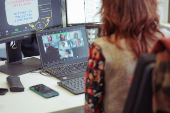 Eine Person sitzt vor einem Laptop und einem großen Bildschirm. Sie ist in einer Videokonferenz mit anderen Menschen, die auf dem Laptop zu sehen sind.