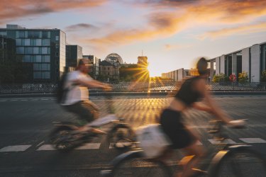 Zwei Personen fahren mit dem Fahrrad, im urbanen Hintergrund geht die Sonne unter.