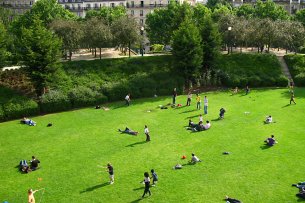 Das Bild zeigt eine grüne Parkfläche an einem sonnigen Tag, auf der sich viele Menschen entspannen und Freizeitaktivitäten nachgehen. Einige Menschen liegen auf dem Gras, während andere Spiele spielen oder miteinander reden. Im Hintergrund sind mehrere mehrstöckige Gebäude zu sehen, die den Park umgeben. Die Szene wirkt lebendig und friedlich, mit viel Grün und einer entspannten Atmosphäre.