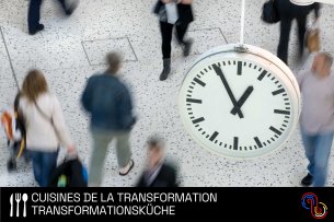 Personen in einem Bahnhof, von oben gesehen. Im Vordergrund eine Uhr.